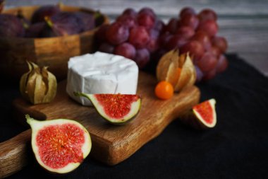 Tahtadaki Brie peynirinin yanında olgun incirler, pembe üzümler ve arka planda Physalis meyveleri.