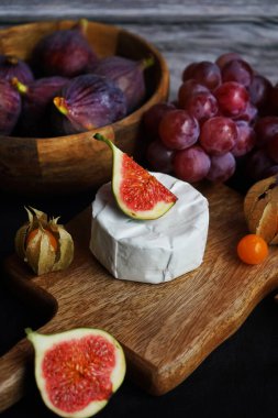 Tahtadaki Brie peynirinin yanında olgun incirler, pembe üzümler ve arka planda Physalis meyveleri.