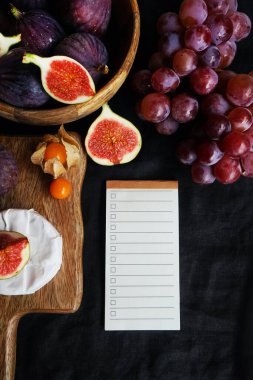 Tahta bir tahtada Brie peynirinin yanında, tahta bir kasede olgun incirler ve karanlık bir arka planda yapılacaklar listesi.