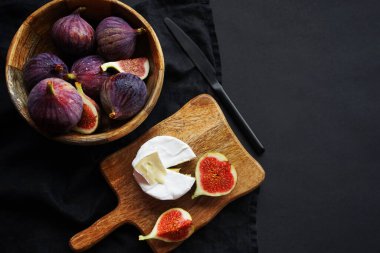 Tahta bir tahtanın üzerinde, tahta bir kasede, Brie peynirinin yanında, koyu bir arka planda olgun incirler.