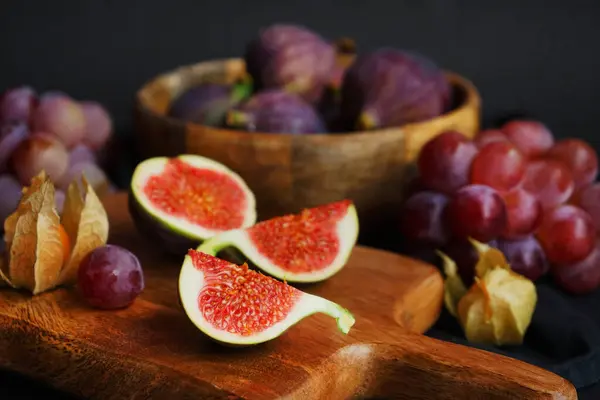 Pembe üzümlerin yanında olgun ve dilimlenmiş incirler ve arka planda tahta bir tahtanın üzerinde Physalis meyveleri.
