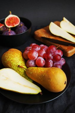 Olgun ve dilimlenmiş incirler, armutlar ve pembe üzümler tahtanın üzerinde ve siyah bir arka planda siyah plaka.