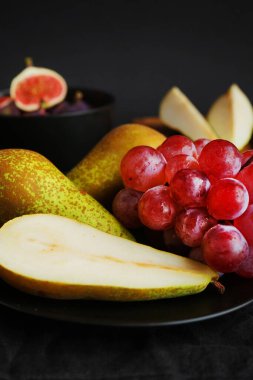 Olgun ve dilimlenmiş incirler, armutlar ve pembe üzümler tahtanın üzerinde ve siyah bir arka planda siyah plaka.