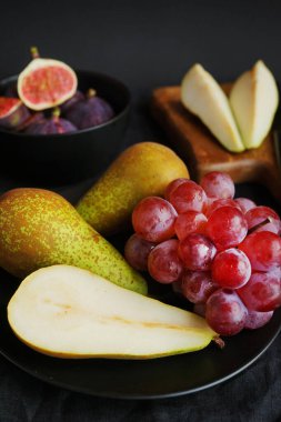 Olgun ve dilimlenmiş incirler, armutlar ve pembe üzümler tahtanın üzerinde ve siyah bir arka planda siyah plaka.
