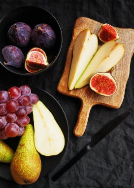 Olgun ve dilimlenmiş incirler, armutlar ve pembe üzümler tahtanın üzerinde ve siyah bir arka planda siyah plaka.