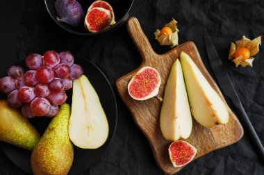 Olgun ve dilimlenmiş incir, armut ve pembe üzümler tahtanın üzerinde ve siyah tabağın yanında karanlık bir arka planda physalis meyveleri.