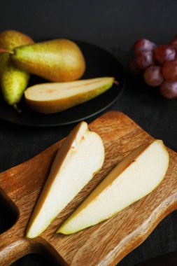 Olgun ve dilimlenmiş armutlar ahşap bir tahta ve tabakta, koyu arkaplanda pembe üzümlerin yanında.