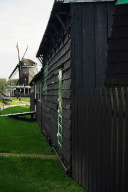 An old wooden house with black walls and green windows against the backdrop of a mill in gloomy weather clipart