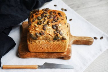 Tahtada balkabağı tohumlu bir somun taze ekmek ve gri arka planda bıçağın yanında pamuk havlu.