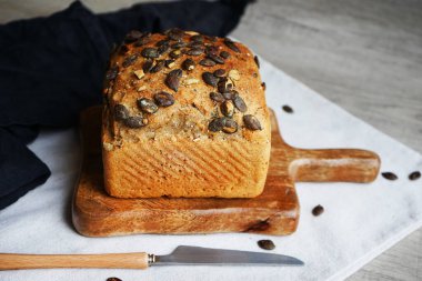 Tahtada balkabağı tohumlu bir somun taze ekmek ve gri arka planda bıçağın yanında pamuk havlu.