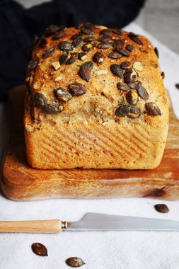 Tahtada balkabağı tohumlu bir somun taze ekmek ve gri arka planda bıçağın yanında pamuk havlu.