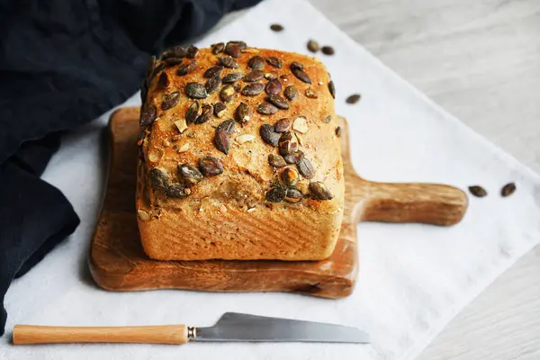 Tahtada balkabağı tohumlu bir somun taze ekmek ve gri arka planda bıçağın yanında pamuk havlu.
