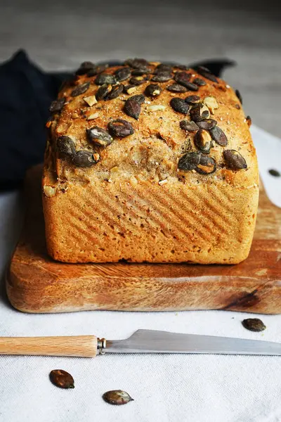 Tahtada balkabağı tohumlu bir somun taze ekmek ve gri arka planda bıçağın yanında pamuk havlu.