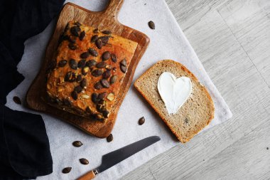 Bir dilim ekmek, tereyağlı kalp, bir somun ekmeğin yanında tahta tahtada kabak çekirdeği ve gri zemin üzerinde bıçak.