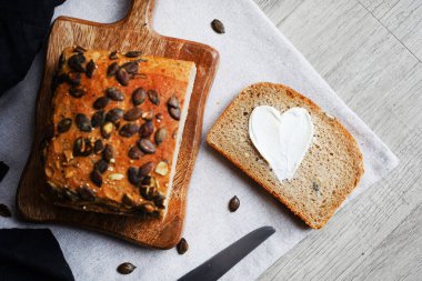 Bir dilim ekmek, tereyağlı kalp, bir somun ekmeğin yanında tahta tahtada kabak çekirdeği ve gri zemin üzerinde bıçak.