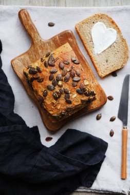 Bir dilim ekmek, tereyağlı kalp, bir somun ekmeğin yanında tahta tahtada kabak çekirdeği ve gri zemin üzerinde bıçak.