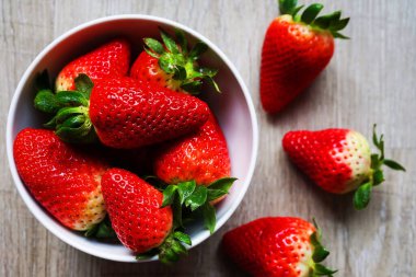 Gri ahşap bir masada çileklerin yanında beyaz bir tabakta büyük olgun çilekler.