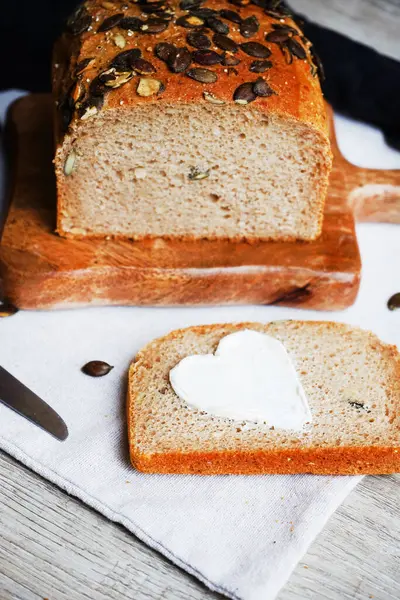 Bir dilim ekmek, tereyağlı kalp, bir somun ekmeğin yanında tahta tahtada kabak çekirdeği ve gri zemin üzerinde bıçak.