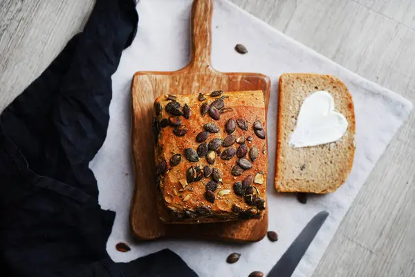 Bir dilim ekmek, tereyağlı kalp, bir somun ekmeğin yanında tahta tahtada kabak çekirdeği ve gri zemin üzerinde bıçak.