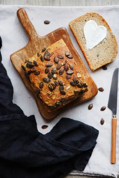 Bir dilim ekmek, tereyağlı kalp, bir somun ekmeğin yanında tahta tahtada kabak çekirdeği ve gri zemin üzerinde bıçak.