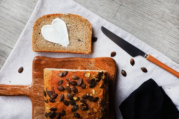 Bir dilim ekmek, tereyağlı kalp, bir somun ekmeğin yanında tahta tahtada kabak çekirdeği ve gri zemin üzerinde bıçak.