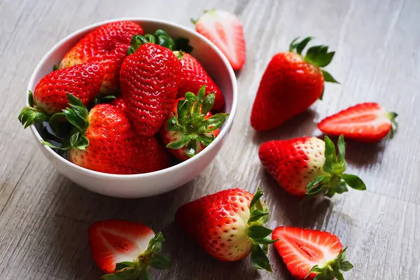 Gri ahşap bir masada çileklerin yanında beyaz bir tabakta büyük olgun çilekler.