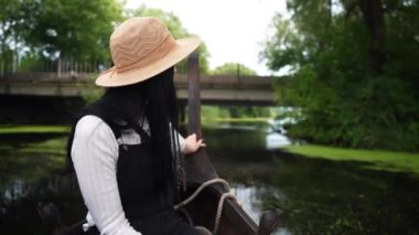 Şapkalı bir kız köprünün önündeki nehirde yelken açan bir teknede oturur ve manzaraya bakar.