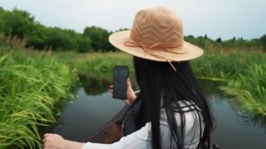 Şapkalı bir kız nehrin taşkın ovasında yüzen bir teknede oturuyor ve telefonuyla manzaranın fotoğrafını çekiyor.