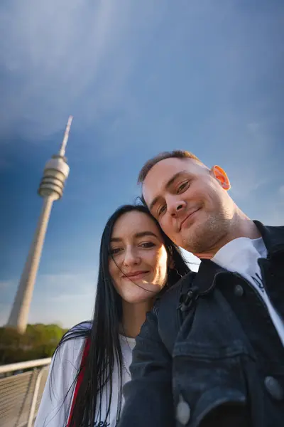 Arka planda mavi gökyüzünün altında uzun bir kuleyle selfie çeken çift.