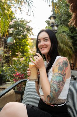 Kafede soğuk kahvenin tadını çıkarırken gülümseyen dövmeli kadın.