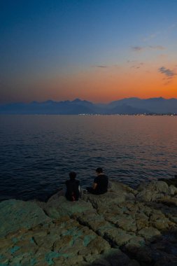 Alacakaranlıkta bir dağın siluetine karşı denizi izleyen iki figür..