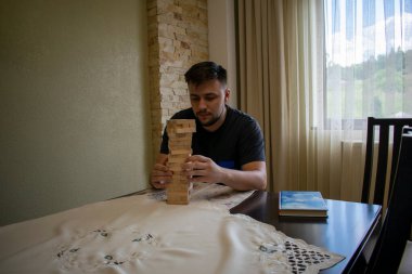 Siyah tişörtlü bir adam masadaki ahşap bir binadan çubuk çekiyor.