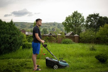 Mavi şortlu ve siyah tişörtlü bir adam, çim biçme makinesinin yardımıyla yeşil çimleri biçiyor. Kırmızı bir taşıyıcıya bağlı, ağaçların ve çitlerin arkasında.