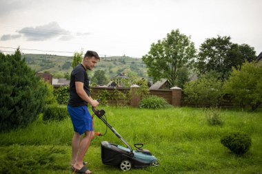 Mavi şortlu ve siyah tişörtlü bir adam, çim biçme makinesinin yardımıyla yeşil çimleri biçiyor. Kırmızı bir taşıyıcıya bağlı, ağaçların ve çitlerin arkasında.