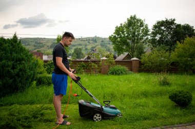 Mavi şortlu ve siyah tişörtlü bir adam, çim biçme makinesinin yardımıyla yeşil çimleri biçiyor. Kırmızı bir taşıyıcıya bağlı, ağaçların ve çitlerin arkasında.