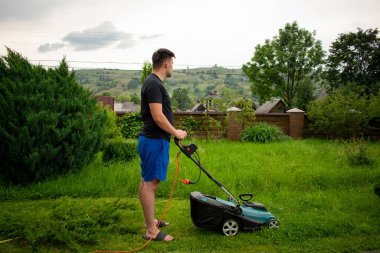 Mavi şortlu ve siyah tişörtlü bir adam, çim biçme makinesinin yardımıyla yeşil çimleri biçiyor. Kırmızı bir taşıyıcıya bağlı, ağaçların ve çitlerin arkasında.