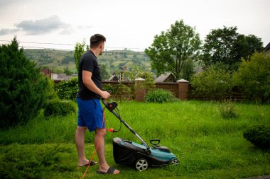 Mavi şortlu ve siyah tişörtlü bir adam, çim biçme makinesinin yardımıyla yeşil çimleri biçiyor. Kırmızı bir taşıyıcıya bağlı, ağaçların ve çitlerin arkasında.