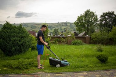 Mavi şortlu ve siyah tişörtlü bir adam, arka plandaki kırmızı bir taşıyıcıya bağlı bir çim biçme makinesi ve kaldırım taşının yanındaki bir çitle yeşil çim biçiyor.
