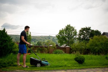 Mavi şortlu ve siyah tişörtlü bir adam, arka plandaki kırmızı bir taşıyıcıya bağlı bir çim biçme makinesi ve kaldırım taşının yanındaki bir çitle yeşil çim biçiyor.
