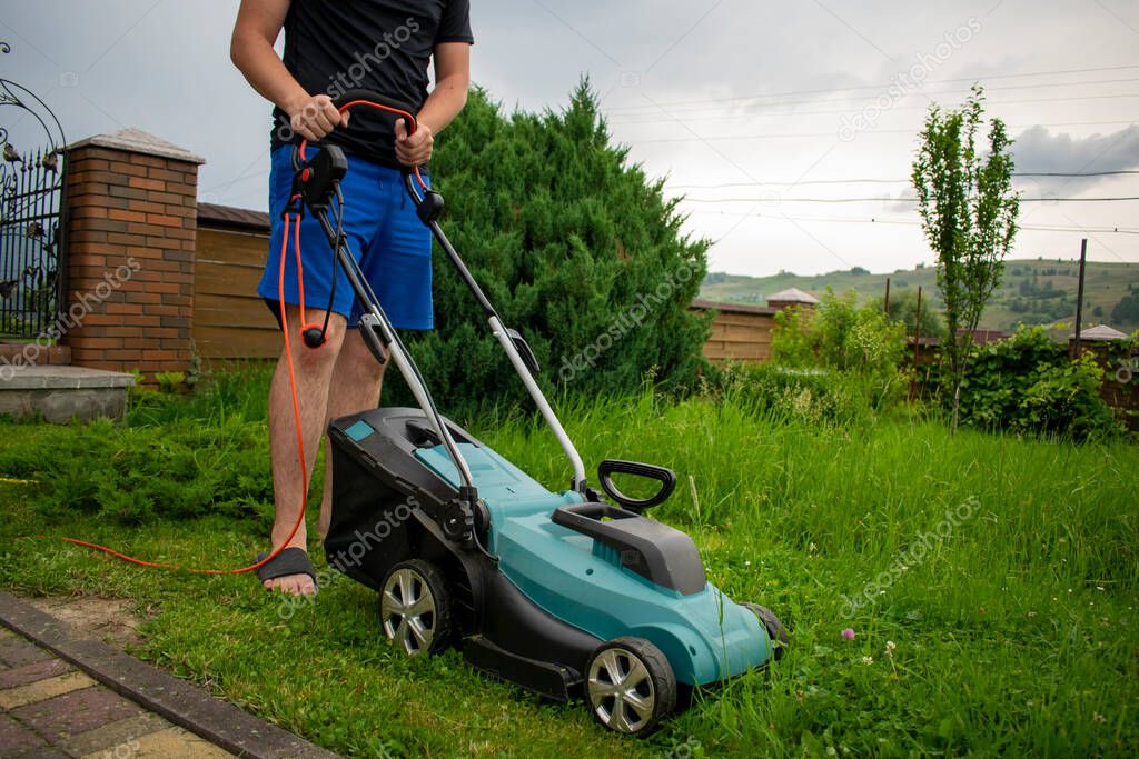 A man in blue shorts and a black t-shirt mows green grass with the help of a lawnmower connected to a red carrier against the background of trees and a fence, holding the lawnmower by the handle