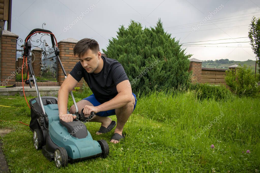 A man in blue shorts and a black T-shirt inspects the condition of a lawnmower in blue and black colors against a background of trees and a fence