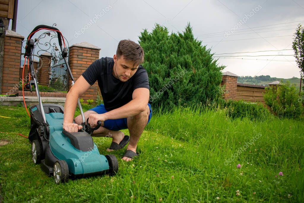 A man in blue shorts and a black T-shirt inspects the condition of a lawnmower in blue and black colors against a background of trees and a fence