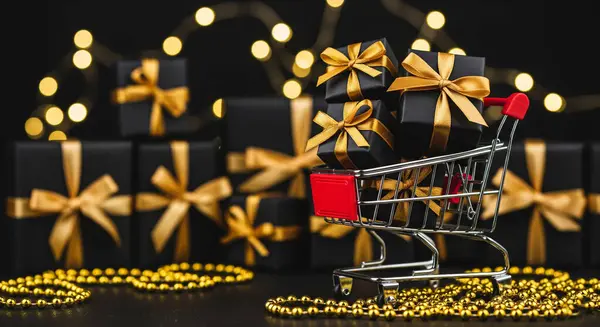 Minyatür bir alışveriş arabası, altın kurdelelerle bağlanmış siyah hediye kutularıyla dolup taşıyor. Arkaplanda bulanık ışık ve altın boncuklar var..