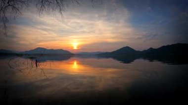 Zaman atlaması, turuncu renkler bulutları boyarken ve dağların sakin sularını yansıtırken gökyüzündeki büyüleyici günbatımını yakalar ve nefes kesici bir manzara yaratır..