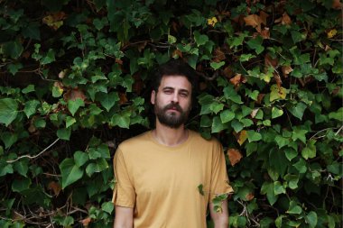 Yeşil yapraklı sarı tişörtlü, sakallı yakışıklı bir genç.