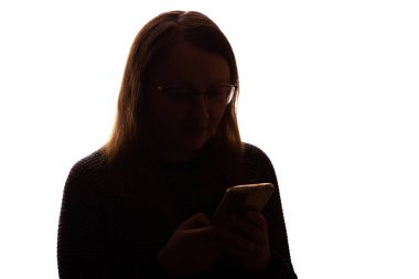 Akıllı telefon tutan ve ekrana bakan genç bir kadının silueti. Beyaz bir arka planda izole edilmiş. Sahne modern iletişim, teknoloji, yalnızlık ve kişisel yansıma temalarını çağrıştırıyor. Temiz ve minimalist kompozisyon.