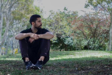 Sporcu kıyafeti giyen genç adam parkta oturmuş yoga yapıyor.