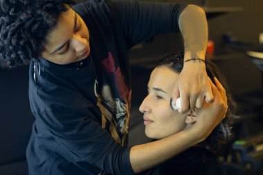 Afro makyöz kadın soyunma odasında genç bir mankene makyaj yapıyor.