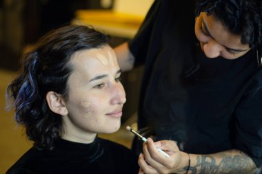 Afro makyöz kadın soyunma odasında genç bir mankene makyaj yapıyor.