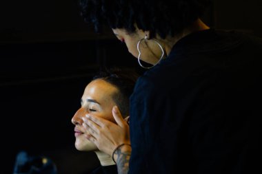 Afro makyöz kadın soyunma odasında genç bir mankene makyaj yapıyor.